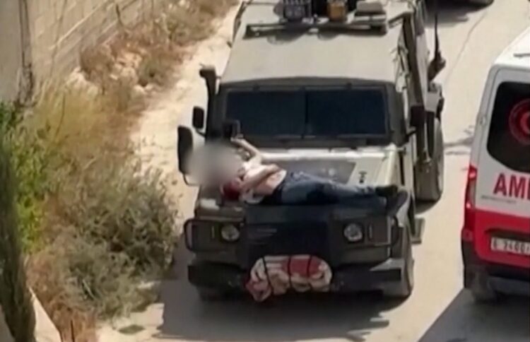 Στο στιγμιότυπο του βίντεο διακρίνεται καθαρά ο τραυματισμένος άνδρας δεμένος πάνω στο καπό του στρατιωτικού τζιπ. Δίπλα του περνά ένα ασθενοφόρο