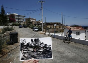 Ο φωτορεπόρτερ κρατάει φωτογραφία, που τραβήχτηκε μια ημέρα μετά την πυρκαγιά και δείχνει μια γυναίκα να περπατάει μπροστά από καμένα αυτοκίνητα, στο Μάτι, ενώ ένα αγόρι περπατάει με το ποδήλατό του στο ίδιο σημείο, στο παραθαλάσσιο θέρετρο, έναν χρόνο μετά (φωτ.: ΑΠΕ-ΜΠΕ/Γιάννης Κολεσίδης)