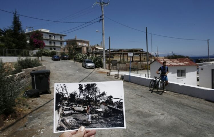 Ο φωτορεπόρτερ κρατάει φωτογραφία, που τραβήχτηκε μια ημέρα μετά την πυρκαγιά και δείχνει μια γυναίκα να περπατάει μπροστά από καμένα αυτοκίνητα, στο Μάτι, ενώ ένα αγόρι περπατάει με το ποδήλατό του στο ίδιο σημείο, στο παραθαλάσσιο θέρετρο, έναν χρόνο μετά (φωτ.: ΑΠΕ-ΜΠΕ/Γιάννης Κολεσίδης)