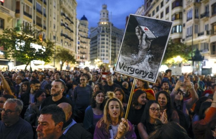 Ισπανίδα στη διάρκεια διαδήλωσης στη Βαλένθια κρατά πανό που γράφει «Ντροπή» και δείχνει ένα χέρι βυθισμένο στα νερά να κρατά ένα κινητό με το μήνυμα έκτακτης ανάγκης, το οποίο άργησε πολύ να φτάσει στους πολίτες (φωτ.: EPA/Kai Forsterling)